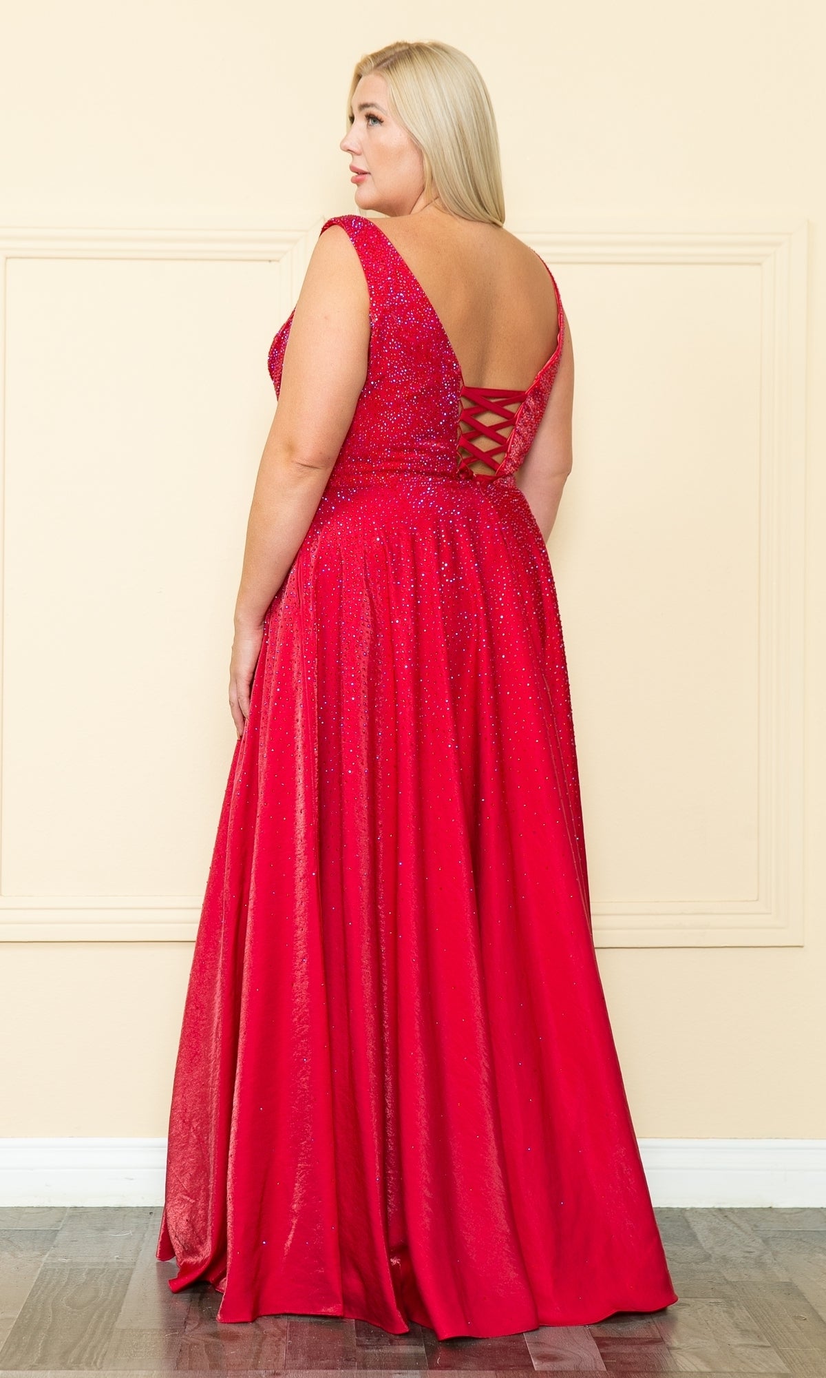 A woman wearing the Embellished Plus-Size Long V-Neck Prom Dress W1110—a sleeveless, floor-length red gown with glitter accents and a lace-up back—stands indoors facing away from the camera.